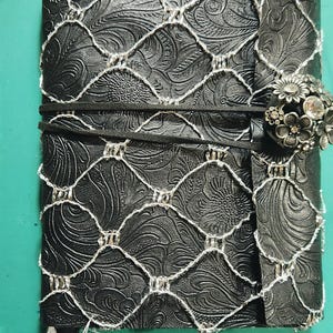 May include: Black leather journal with embossed floral pattern, overlaid with a white stitched net design. The journal is secured with a black elastic band and a decorative silver clasp with floral and rhinestone accents.