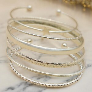 Peut inclure: Un ensemble de sept bracelets argentés, empilés les uns sur les autres. Les bracelets sont de différentes largeurs et ont des textures variées, notamment lisses, texturées et perlées.