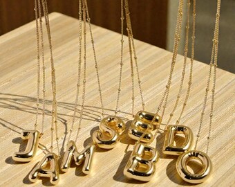 May include: Gold-colored initial necklaces. Each necklace features a different letter pendant, including J, A, M, S, E, B, D, and O. The pendants are suspended from delicate gold chains. The necklaces are displayed on a light-colored wooden surface.