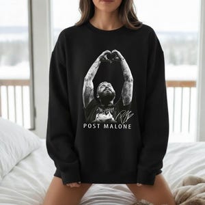 May include: Black sweatshirt featuring a black and white image of Post Malone with his arms raised in a heart shape. The text "POST MALONE" and a signature are printed below the image. The sweatshirt is a crew neck style.