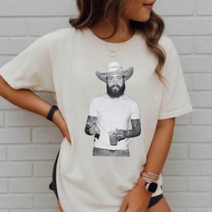 May include: A cream-colored short-sleeve t-shirt featuring a black and white graphic print of a bearded person wearing a cowboy hat, a plain t-shirt, and holding a beverage. The graphic is centered on the chest of the garment.
