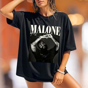 May include: A black short-sleeved t-shirt featuring a large graphic print on the front. The graphic displays a black and white photograph of a bearded person making a heart shape with their hands, with the text MALONE POST printed above.