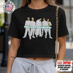 May include: Black crop top featuring a group of men in white outfits with the text "MILLENNIUM 20" in a light blue font. The top has a crew neck and short sleeves. A "Made in the USA" sticker is visible.