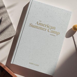 Op de afbeelding: Een wit dagboek met de tekst "The American Summer Camp Journal" in gouden letters. Het dagboek ligt op een lichtbeige oppervlak.