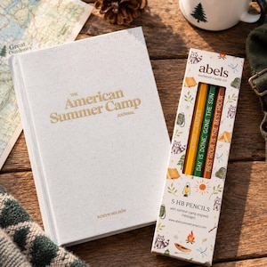 May include: A white hardback journal titled The American Summer Camp Journal by Robyn Wilson, alongside a rectangular box containing five HB pencils. The pencil box features illustrations of raccoons, tents, and camp gear with text.