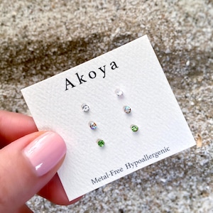 May include: A white card with the word "Aikoa" printed in black. The card has six small, clear, round rhinestones arranged in two rows of three. The card also has the text "Metal-Free Hypoallergenic" printed in black.