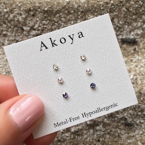 May include: A set of three pairs of small, clear crystal stud earrings on a white card with the text "Aikoa Metal-Free Hypoallergenic".