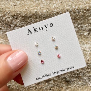 Peut inclure: Une carte blanche avec le mot "Aikoa" imprimé en noir. La carte comporte cinq petits strass ronds et transparents disposés en motif. Le texte "Metal-Free Hypoallergenic" est imprimé en bas de la carte.