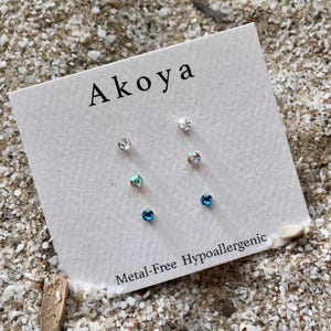 May include: A set of six small stud earrings on a white card with the text "Aikoa Metal-Free Hypoallergenic". The earrings are silver with clear, blue, and iridescent stones.