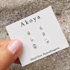 Peut inclure: Une carte blanche avec le mot "Aikoa" imprimé en noir. La carte présente cinq boucles d'oreilles en cristal transparent. Le texte "Metal-Free Hypoallergenic" est imprimé en bas de la carte.