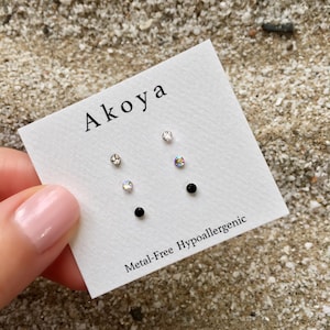 May include: A white card with the word "Aikoa" printed in black. The card has three pairs of earrings on it: one pair of clear crystal earrings, one pair of iridescent crystal earrings, and one pair of black stud earrings. The card also says "Metal-Free Hypoallergenic".