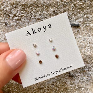 Peut inclure: Un ensemble de six petites boucles d'oreilles à clous en cristal transparent et iridescent sur une carte blanche avec le texte "Akoya" et "Metal-Free Hypoallergenic".
