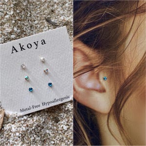 May include: A set of three small stud earrings with clear and blue crystals. The earrings are on a white card with the brand name "Aikoa" and the text "Metal-Free Hypoallergenic".