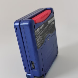 May include: A blue Game Boy Advance SP handheld game console with a red power button. The console is angled, showing the back with text and a power switch. The device is a classic retro gaming console.
