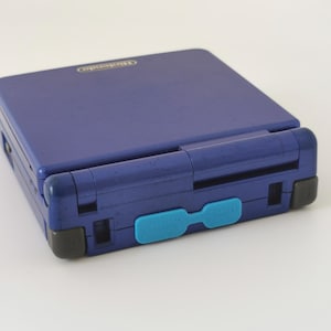 Könnte beinhalten: Eine violette Nintendo Game Boy Advance SP Handheld-Spielkonsole. Die Konsole ist geschlossen und zeigt die Oberseite und das Scharnier. Der Power-Knopf ist schwarz und die Abdeckung des Spielkartensteckplatzes hellblau. Das Nintendo-Logo ist sichtbar.