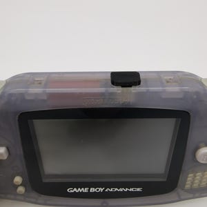 May include: A translucent gray Game Boy Advance handheld gaming console with a black screen and the text "GAME BOY ADVANCE" printed on the front.