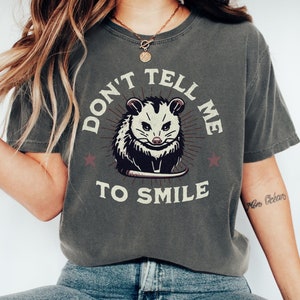 May include: A gray t-shirt with a graphic of a possum with the text "Don't Tell Me To Smile" in a vintage style.