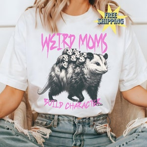 May include: White t-shirt featuring a graphic of an opossum carrying baby opossums on its back. The text "Weird Moms Build Character" is printed in pink below the opossum.