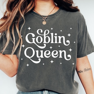 Puede incluir: Una camiseta gris oscuro con un gráfico blanco que dice "Goblin Queen" en una fuente retro con estrellas blancas que rodean el texto.