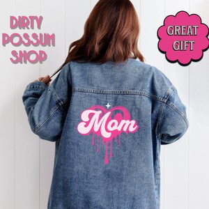 May include: A blue denim jacket with a pink heart design that says "Mom" in a retro font. The heart is dripping with pink paint.