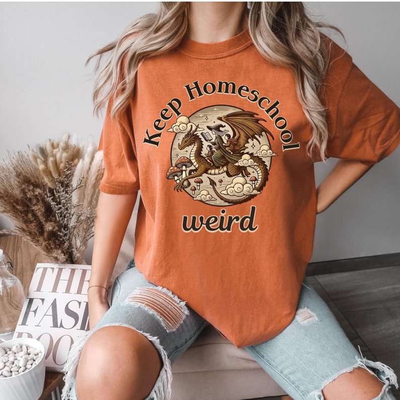 以下が含まれることがあります： 「Keep Homeschool Weird」というテキストと、ドラゴンに乗った魔女のグラフィックが描かれたオレンジ色のTシャツです。