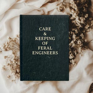 Könnte beinhalten: Ein schwarzes Buch mit dem Titel "Care & Keeping of Feral Engineers" in goldenen Lettern. Das Buch liegt auf einer beigefarbenen Stoffoberfläche mit getrockneten weißen Blumen.
