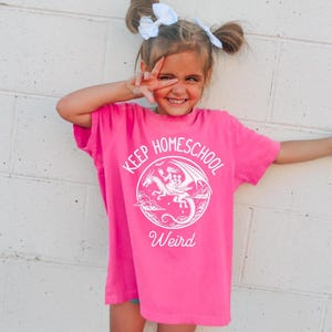 May include: A bright pink t-shirt with the words "Keep Homeschool Weird" in white, featuring a dragon graphic. The shirt is worn by a young person with white bows in their hair.