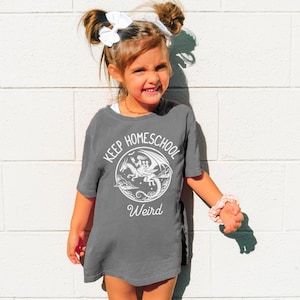 May include: A gray t-shirt with the words "KEEP HOMESCHOOL Weird" in white. The shirt features a circular graphic of a dragon. The girl is wearing a white bow and a pink scrunchie.
