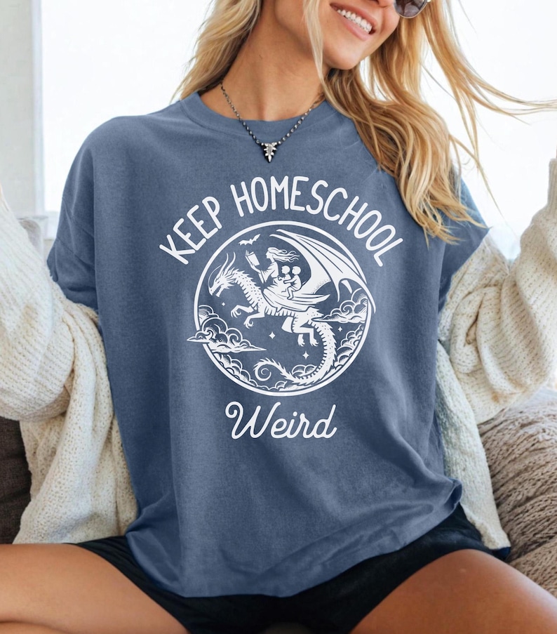 May include: A blue t-shirt with a white graphic of a woman riding a dragon with the text "Keep Homeschool Weird".