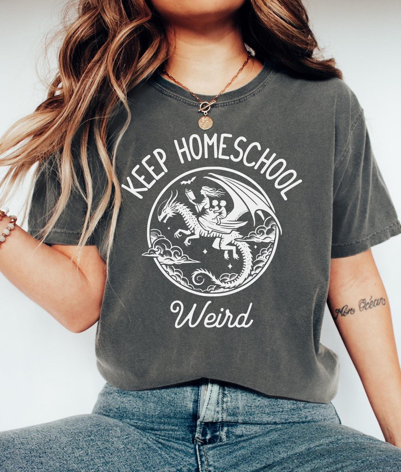 May include: A dark gray t-shirt with a white graphic of a person riding a dragon with the text "Keep Homeschool Weird".
