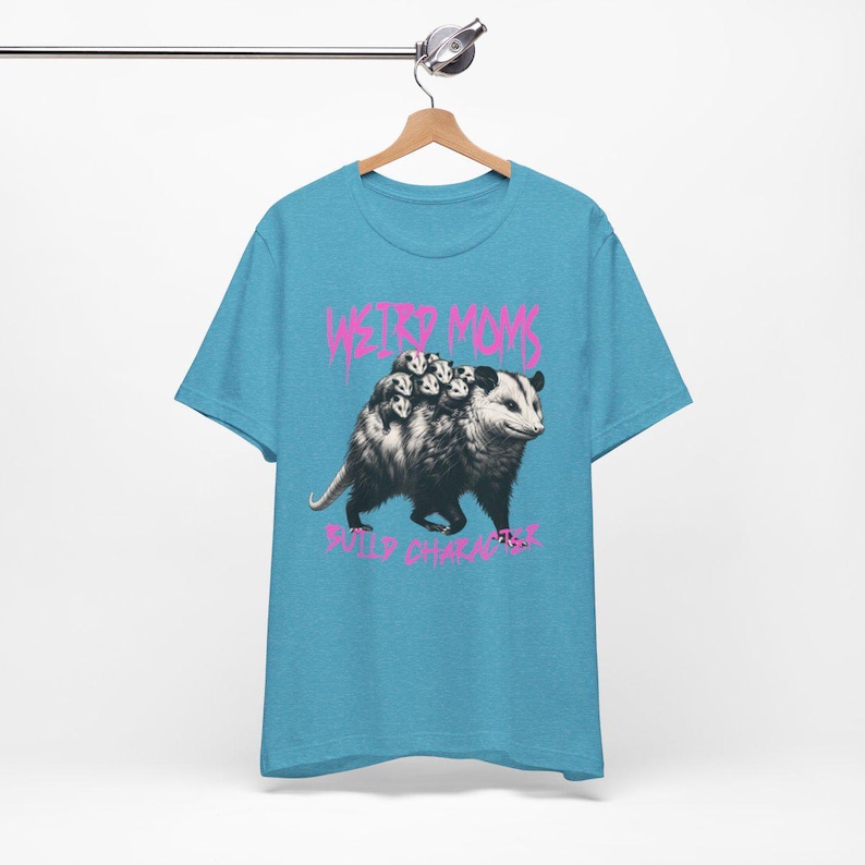 May include: A light blue t-shirt with a graphic of a possum carrying a group of baby possums on its back. The text "Weird Moms Build Character" is printed in pink below the graphic.