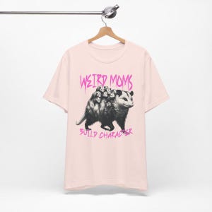 May include: Pink t-shirt with a graphic of a possum carrying baby possums on its back. The text "Weird Moms Build Character" is printed in pink below the image.
