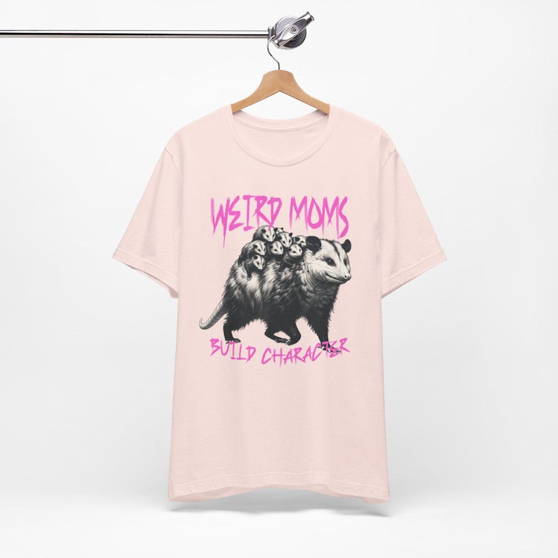 May include: Pink t-shirt with a graphic of a possum carrying baby possums on its back. The text "Weird Moms Build Character" is printed in pink below the image.