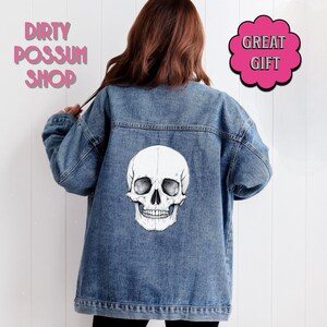 May include: A blue denim jacket with a white skull graphic on the back. The jacket has a relaxed fit and long sleeves. The text "Dirty Possum Shop" is printed in pink on the top left corner of the image. The text "Great Gift" is printed in pink on a white cloud shape in the top right corner of the image.
