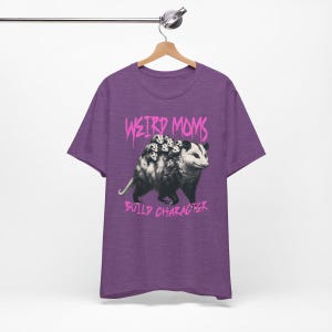 May include: Purple t-shirt with a graphic of a possum carrying baby possums on its back. The text "Weird Moms Build Character" is printed in pink below the image.
