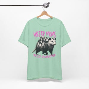 May include: A light green t-shirt with a graphic of a possum carrying baby possums on its back. The text "Weird Moms Build Character" is printed in pink below the image.