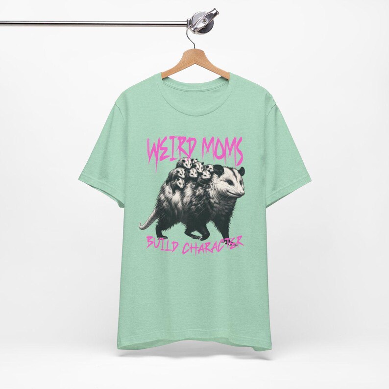 May include: A light green t-shirt with a graphic of a possum carrying baby possums on its back. The text "Weird Moms Build Character" is printed in pink below the image.