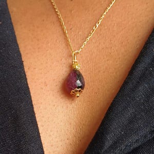 May include: A gold-colored necklace with a teardrop-shaped pendant. The pendant is a mix of dark red and black gemstone, with gold-colored accents. The necklace is worn against a dark blue fabric.