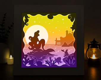 May include: A papercraft light-up scene featuring a mermaid sitting on a rock with a castle in the background. The scene is illuminated with a warm yellow light, creating a magical atmosphere. The words "CRAFT MOON ART" are visible in the top left corner.