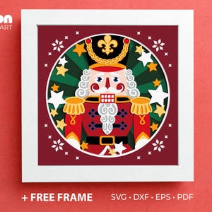 May include: A framed, colorful illustration of a nutcracker soldier. The artwork features a red uniform, white hair, and a stern expression, surrounded by stars and a green and gold background. The frame is white, and the background is red. Text at the bottom reads "+ FREE FRAME".