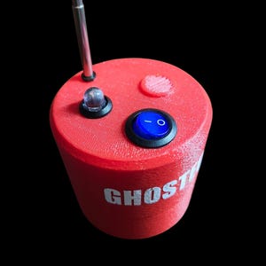 Op de afbeelding: Een cilindervormig rood apparaat met een textuur op het oppervlak en het woord GHOST in witte letters. De bovenkant bevat een zilveren telescopische antenne, een heldere ledlamp en een blauwe tuimelschakelaar met een wit aan-uit symbool.