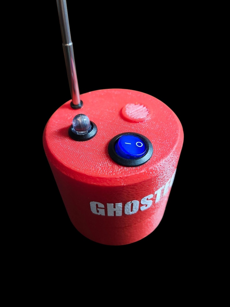 Op de afbeelding: Een cilindervormig rood apparaat met een textuur op het oppervlak en het woord GHOST in witte letters. De bovenkant bevat een zilveren telescopische antenne, een heldere ledlamp en een blauwe tuimelschakelaar met een wit aan-uit symbool.