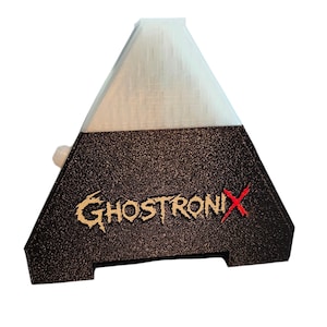 May include: A pyramid-shaped device with a translucent white top section and a textured black base. The word GHOSTRONIX is printed in a stylized font on the front, with the letter X highlighted in red. A small white circular button is visible on the side.
