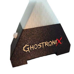 May include: A pyramid-shaped electronic device with a textured black base and a translucent white top section. The front features the text GHOSTRONIX in a stylized font with a red X. A small circular white button is visible on the side panel.