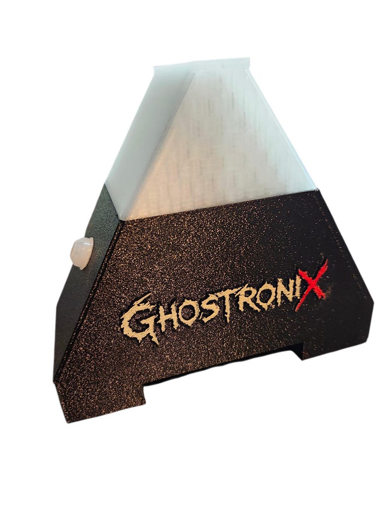 May include: A pyramid-shaped electronic device with a textured black base and a translucent white top section. The front features the text GHOSTRONIX in a stylized font with a red X. A small circular white button is visible on the side panel.