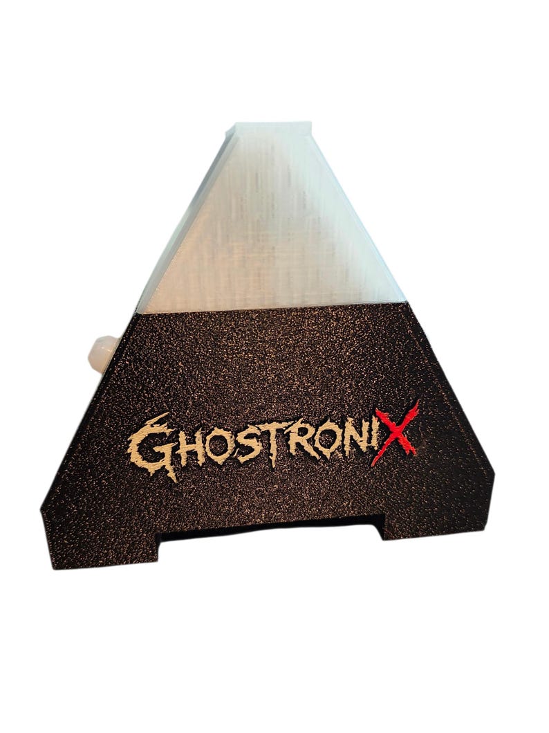 May include: A pyramid-shaped object with a white translucent top section and a textured black base. The text GHOSTRONIX is printed on the front in a stylized font, with the letter X highlighted in red. A small white circular button protrudes from the side.