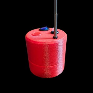 Op de afbeelding: Een cilindervormig rood apparaat met een getextureerd oppervlak, voorzien van een blauwe tuimelschakelaar, een klein rond luidsprekerrooster en een verticale metalen antenne aan de bovenzijde. Het object heeft een naad tussen de boven- en onderhelft.
