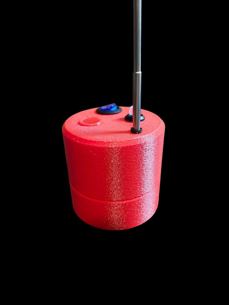 Op de afbeelding: Een cilindervormig rood apparaat met een getextureerd oppervlak, voorzien van een blauwe tuimelschakelaar, een klein rond luidsprekerrooster en een verticale metalen antenne aan de bovenzijde. Het object heeft een naad tussen de boven- en onderhelft.