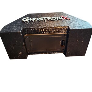 May include: A black, textured, trapezoidal electronic device featuring the word GHOSTRONIX in raised, white, distressed lettering with a red X. The front panel includes a rectangular battery compartment secured by four small black screws.