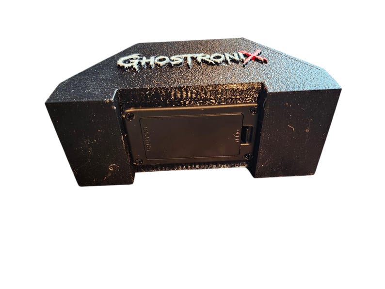 May include: A black, textured, trapezoidal electronic device featuring the word GHOSTRONIX in raised, white, distressed lettering with a red X. The front panel includes a rectangular battery compartment secured by four small black screws.